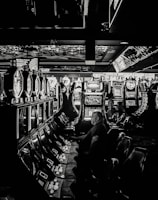 A dimly lit casino filled with rows of slot machines, some occupied by people. The machines' lights contrast with the dark surroundings, creating a vibrant yet subdued atmosphere.