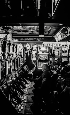 A dimly lit casino filled with rows of slot machines, some occupied by people. The machines' lights contrast with the dark surroundings, creating a vibrant yet subdued atmosphere.