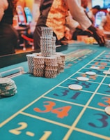 A casino scene with a roulette table features prominently, covered in green felt with colorful numbers and betting squares. Several stacks of poker chips are placed on different numbers, and the hands of several people are visible as they interact with the table. In the background, there are slot machines and more people, creating a vibrant and busy atmosphere.
