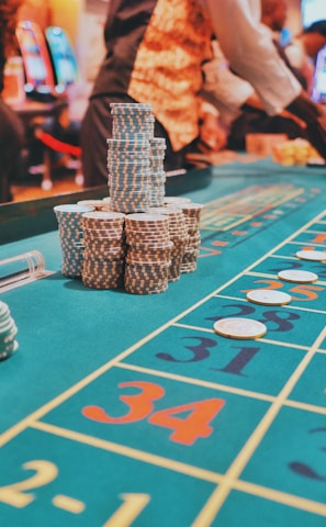 A casino scene with a roulette table features prominently, covered in green felt with colorful numbers and betting squares. Several stacks of poker chips are placed on different numbers, and the hands of several people are visible as they interact with the table. In the background, there are slot machines and more people, creating a vibrant and busy atmosphere.