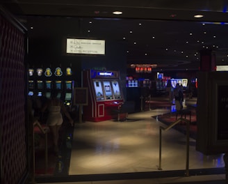 A dimly lit casino with several rows of slot machines, some displaying bright graphics and lights. There are a few people scattered throughout the area, engaging with the machines. The floor is reflective and polished, adding to the ambiance, while the ceiling is dotted with numerous small lights.