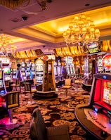 A vibrant casino interior filled with rows of colorful slot machines, each illuminated with bright lights and displays. The ceiling is adorned with elegant chandeliers and golden drapes, creating a luxurious atmosphere. The patterned carpet adds to the elaborate design of the space.
