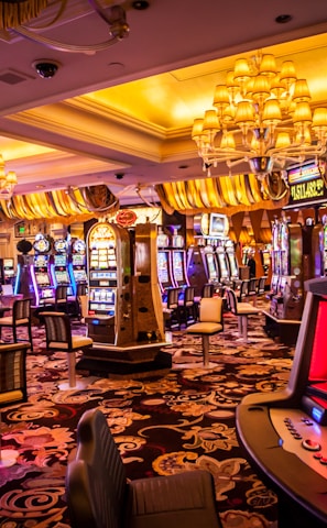A vibrant casino interior filled with rows of colorful slot machines, each illuminated with bright lights and displays. The ceiling is adorned with elegant chandeliers and golden drapes, creating a luxurious atmosphere. The patterned carpet adds to the elaborate design of the space.