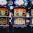 A row of colorful slot machines lit with bright neon lights, showcasing various graphical elements on the screens, indicating different games and jackpots. The atmosphere suggests a lively casino setting, bustling with excitement.