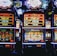 A row of colorful slot machines lit with bright neon lights, showcasing various graphical elements on the screens, indicating different games and jackpots. The atmosphere suggests a lively casino setting, bustling with excitement.