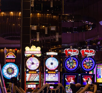 A lively casino scene featuring a row of bright, colorful slot machines with themes like Monopoly, Michael Jackson, and Lord of the Rings. The gaming machines display a variety of flashing lights and digital screens, creating an energetic and exciting atmosphere. Reflections of the lights are visible in glossy surfaces above the machines.