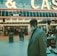 An older man with a gray beard and wearing a suit and hat stands in front of a brightly lit casino. The background features a large illuminated sign reading 'CASINO' and 'SLOTS' among other words, with numerous lights outlining the entrance. Several other people are visible in the background, engaged in various activities, some walking, some standing, and one looking at something on display.