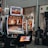 A collection of slot machines is situated inside a building with bright, colorful screens displaying various games and gaming themes. A person is seated nearby, seemingly relaxed with luggage beside them, and the atmosphere is lively with electronic displays.